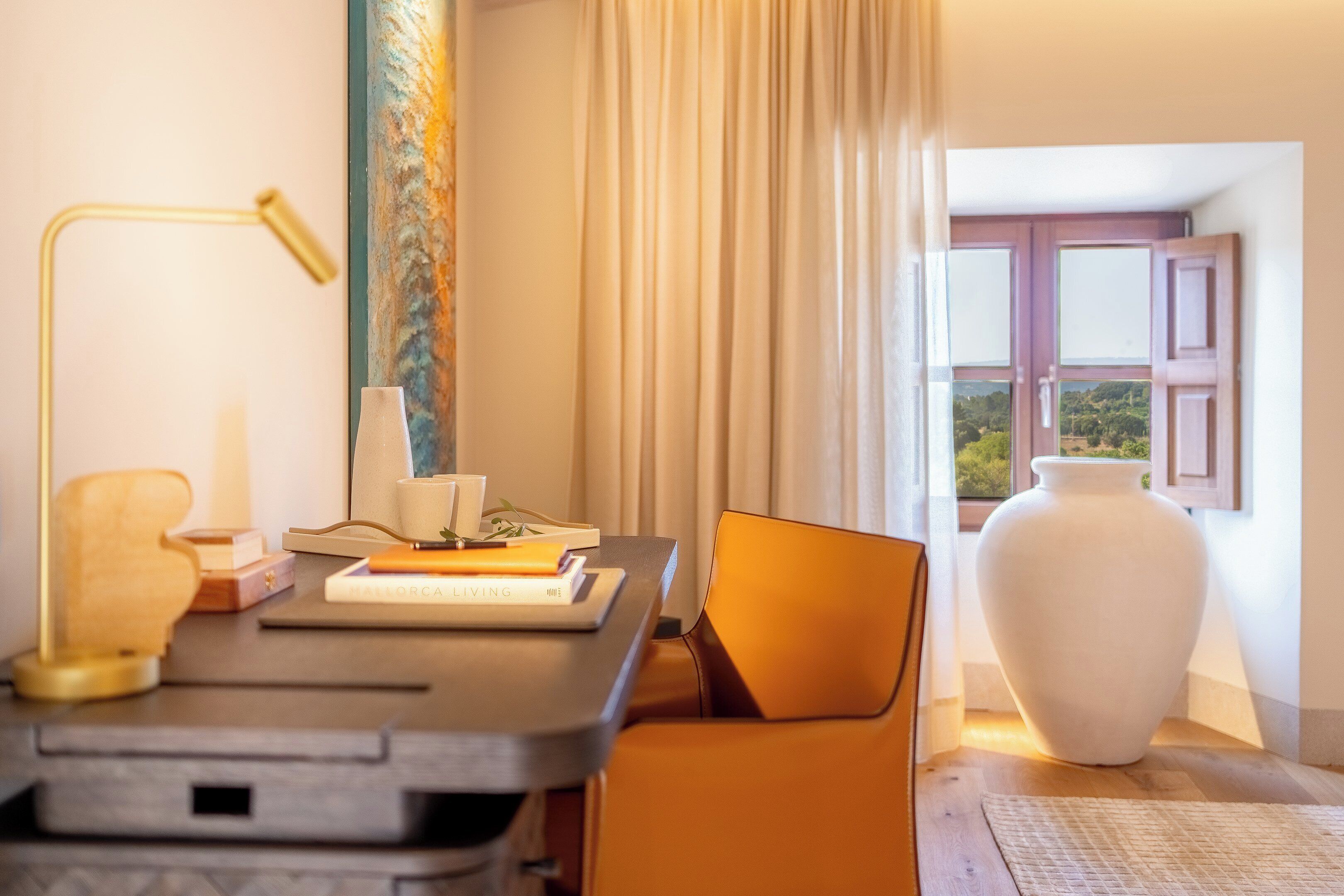 Una habitación con escritorio, lámpara, silla, un jarrón y vistas al exterior.
