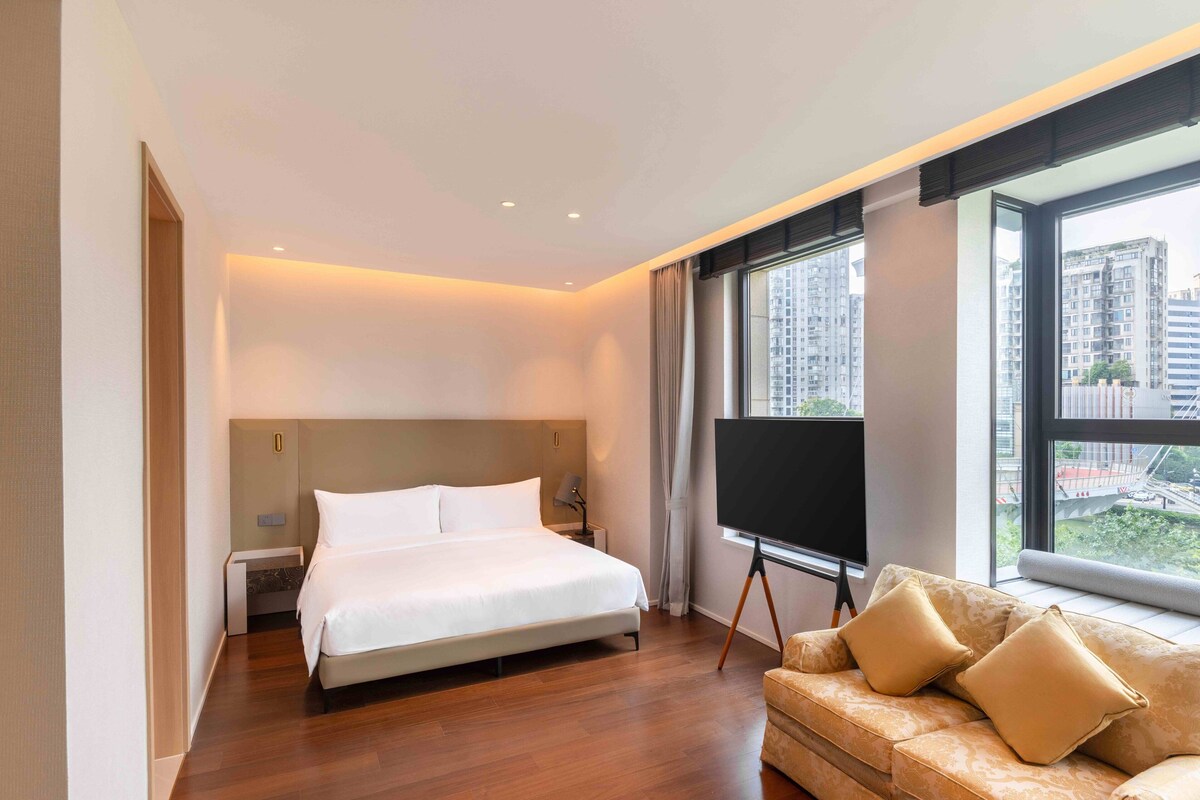 Ein modernes Hotelzimmer mit einem großen Bett, einer Couch, einem Flachbildfernseher und Blick auf hohe Gebäude.