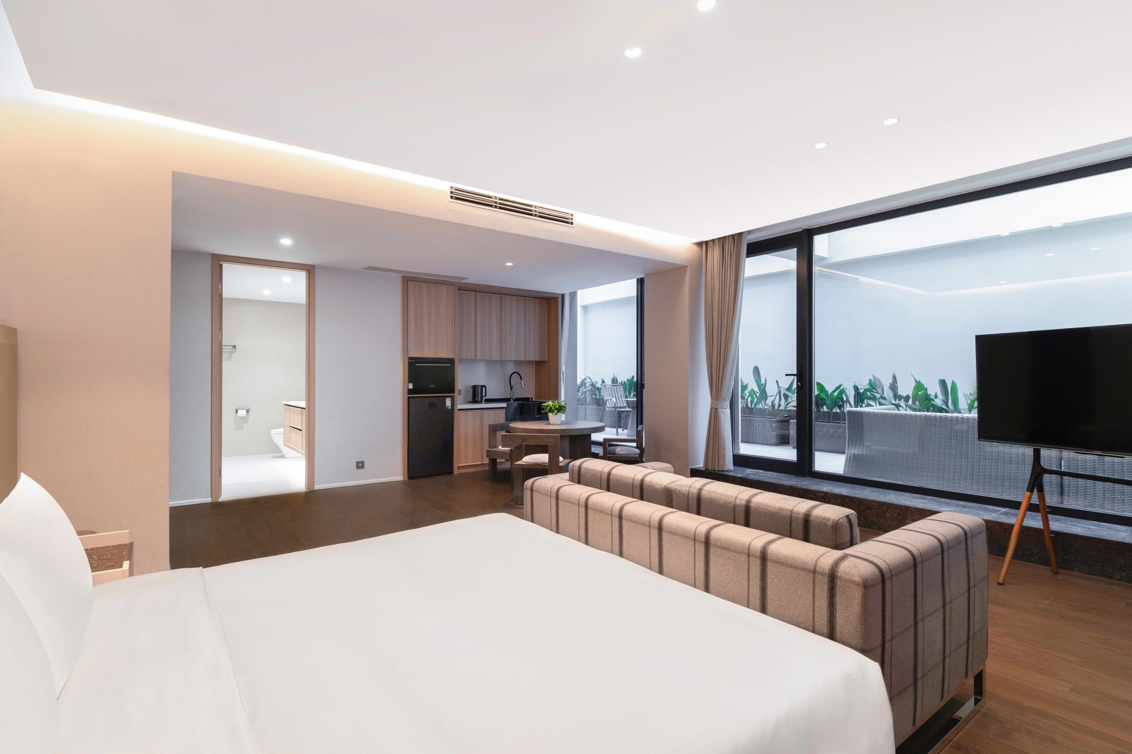 A modern hotel room with a large bed, a seating area, a kitchenette, and a TV.