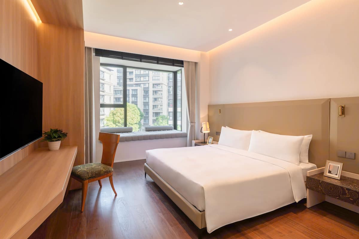 Une chambre d’hôtel moderne dotée d’un grand lit, d’un téléviseur à écran plat, d’une fenêtre donnant sur des immeubles et d’une petite table avec une lampe.