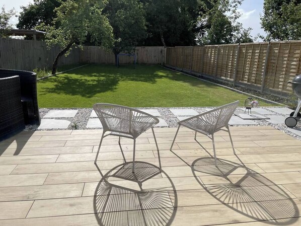 A patio with two metal chairs and a table, surrounded by a wooden deck and a fenced backyard.
