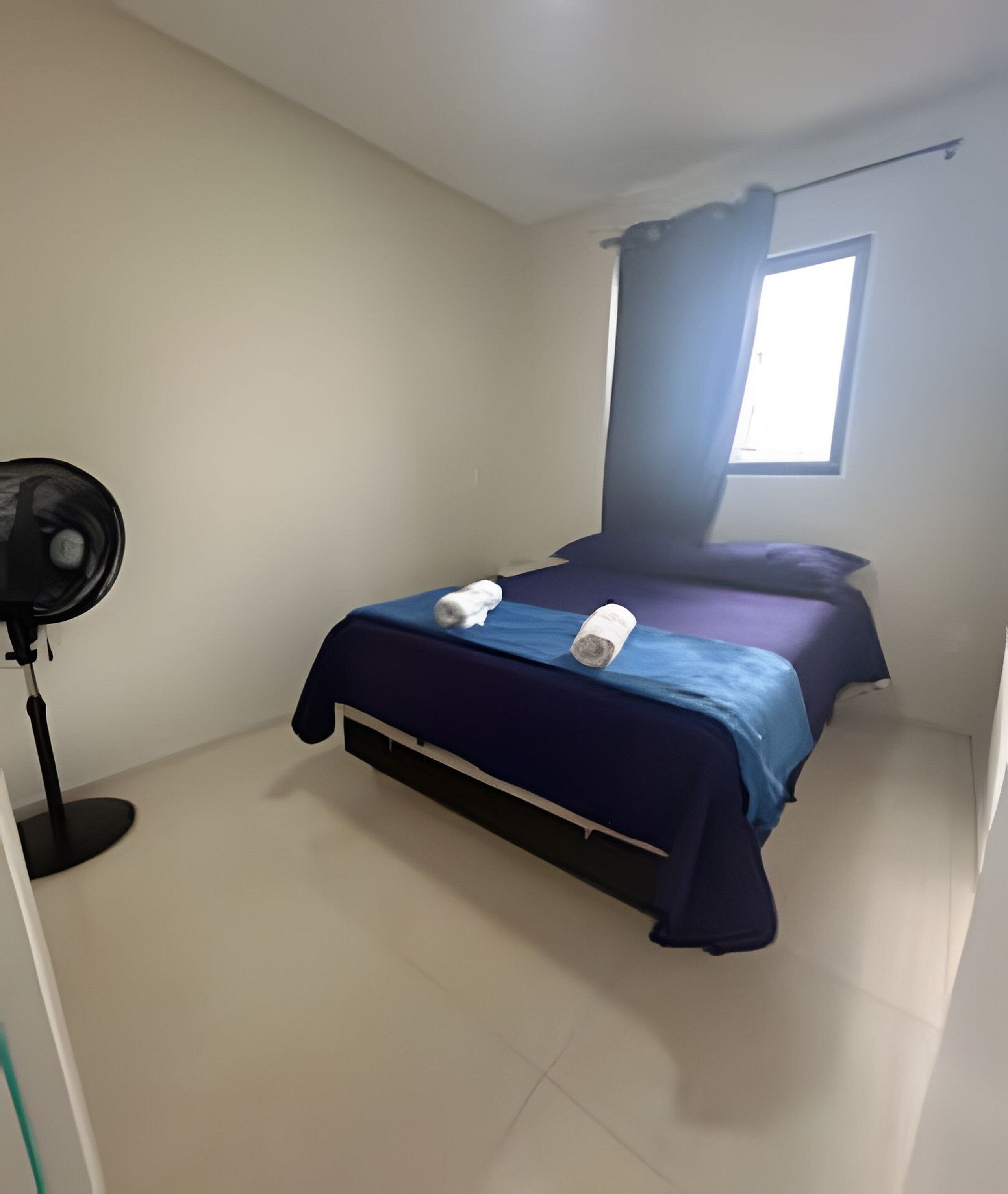 A bedroom with a bed, a standing fan, and a window with curtains.
