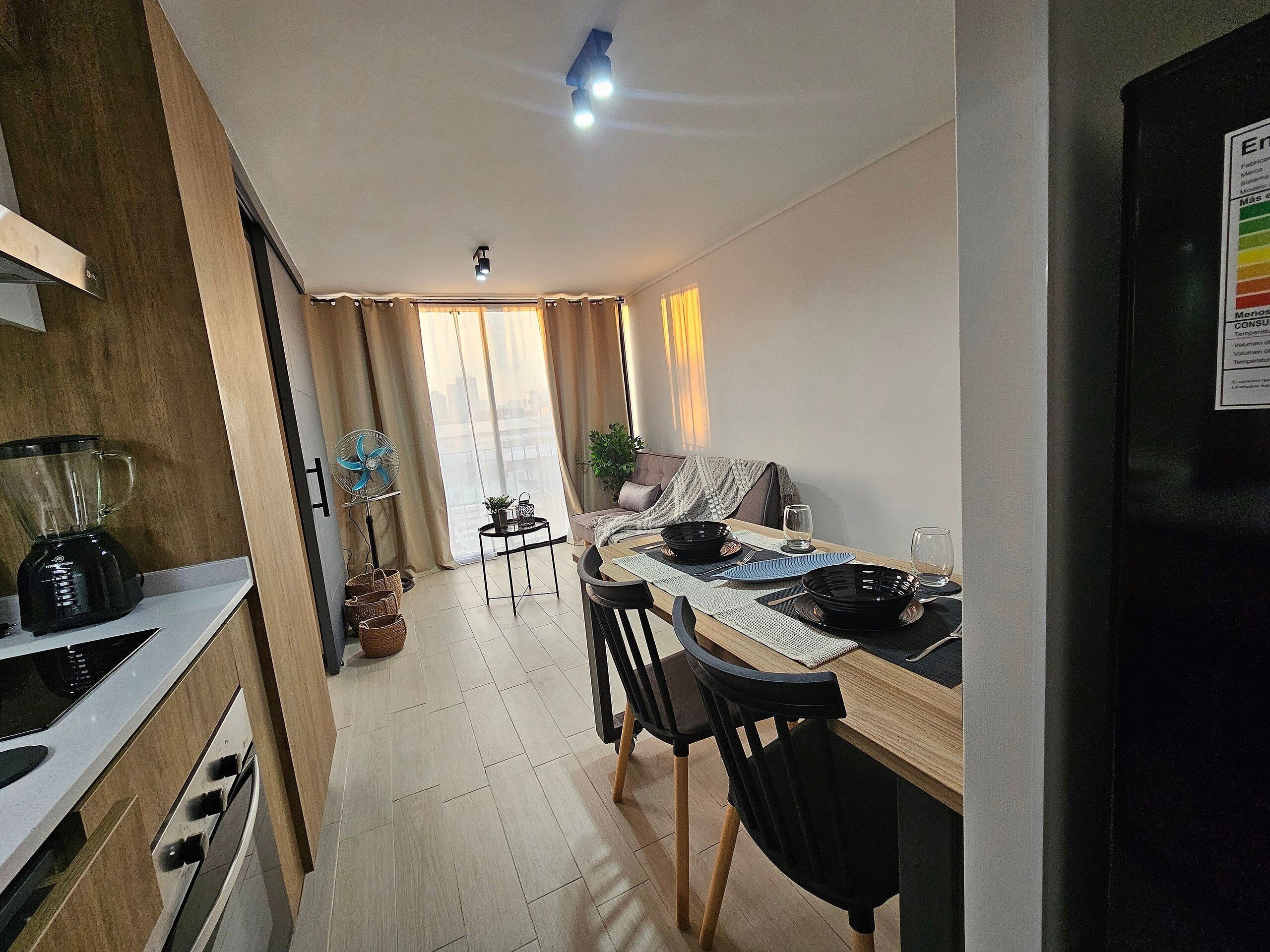 Una cocina moderna con horno empotrado, licuadora y ventilador. Hay una zona de comedor con una mesa para dos, un sofá y una planta.