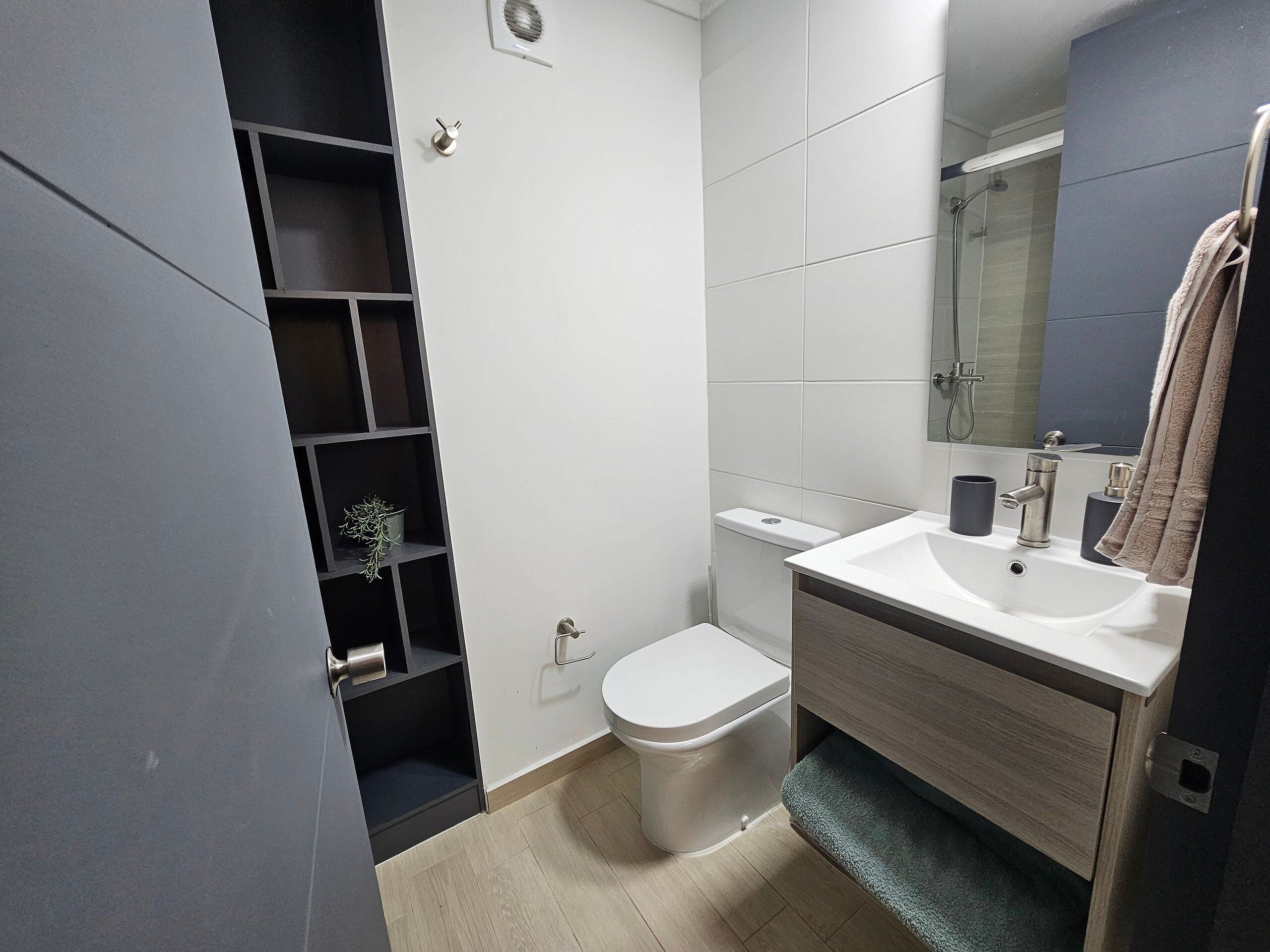 A modern bathroom with a toilet, sink, and mirror. There is a shelf with a plant and a towel hanging on the wall.