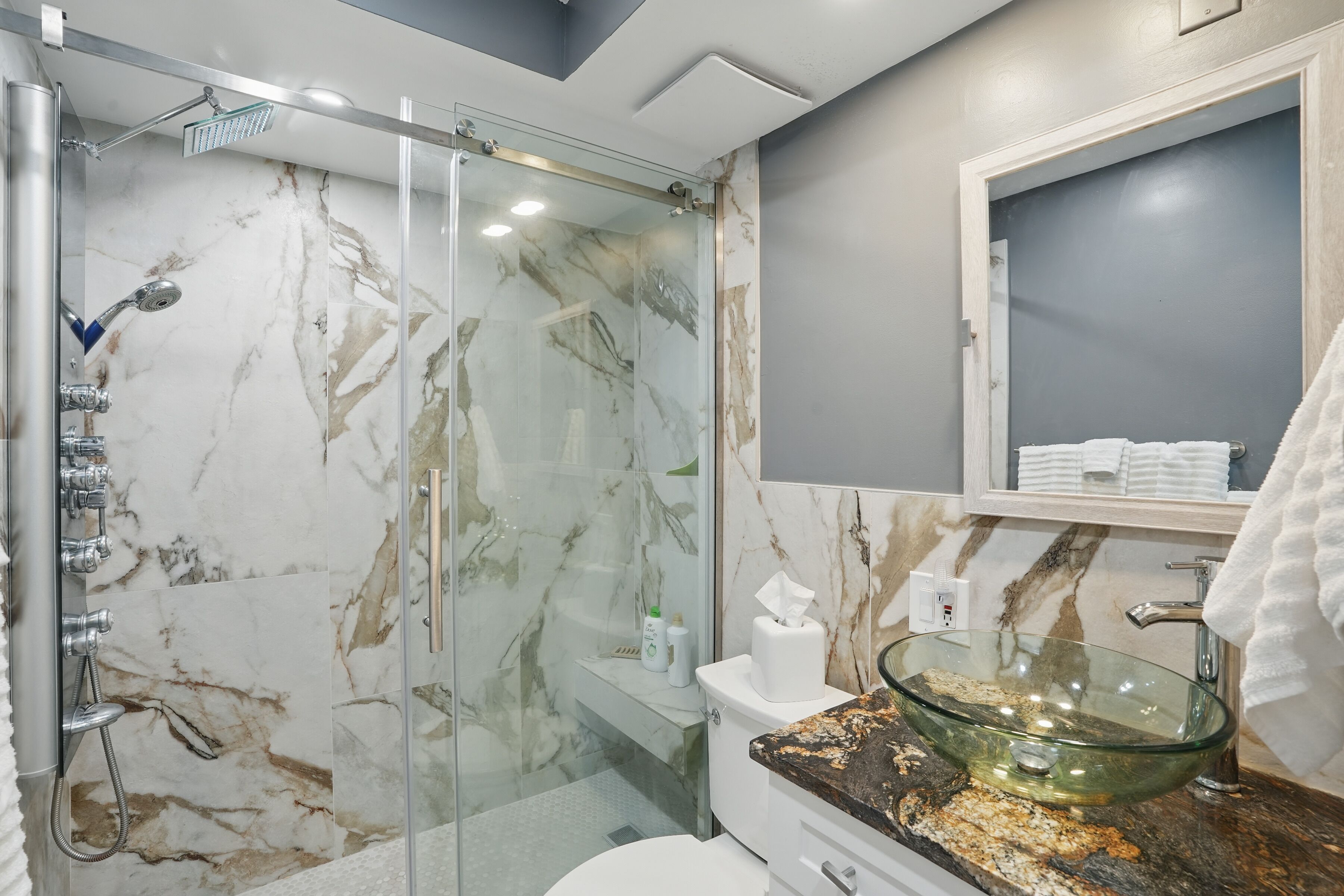 A modern bathroom with a glass-enclosed shower, marble walls, a glass vessel sink, and a white toilet.