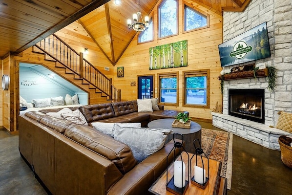 Interior acogedor de una cabaña con una escalera de madera, un sofá de cuero tipo seccional, una chimenea y un ventanal con vista a un lago.