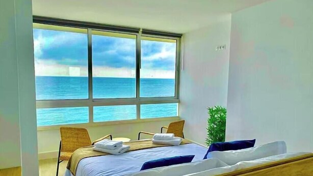 A bedroom with a large window overlooking the sea, a bed with white linens, a wooden headboard, and a small potted plant. - Front line beach Benalmádena La Perla Playa Paraíso (BENALMÁDENA)