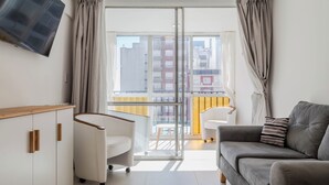 Uma sala de estar moderna com um sofá, uma poltrona branca e um armário de madeira. Uma televisão está fixada na parede, e é visível uma varanda com corrimão amarelo pela porta de vidro.