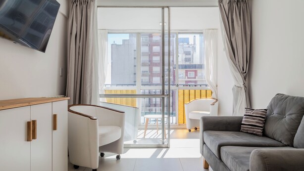A modern living room with a sofa, a white armchair, and a wooden cabinet. A television is mounted on the wall, and a balcony with a yellow railing is visible through the glass door. - BT Homes - Mar del Plata, Av. Colon (Mar del Plata)