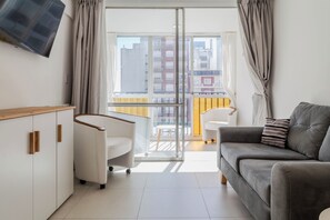 A modern living room with a sofa, a white armchair, and a wooden cabinet. A television is mounted on the wall, and a balcony with a yellow railing is visible through the glass door. - BT Homes - Mar del Plata, Av. Colon (Mar del Plata)