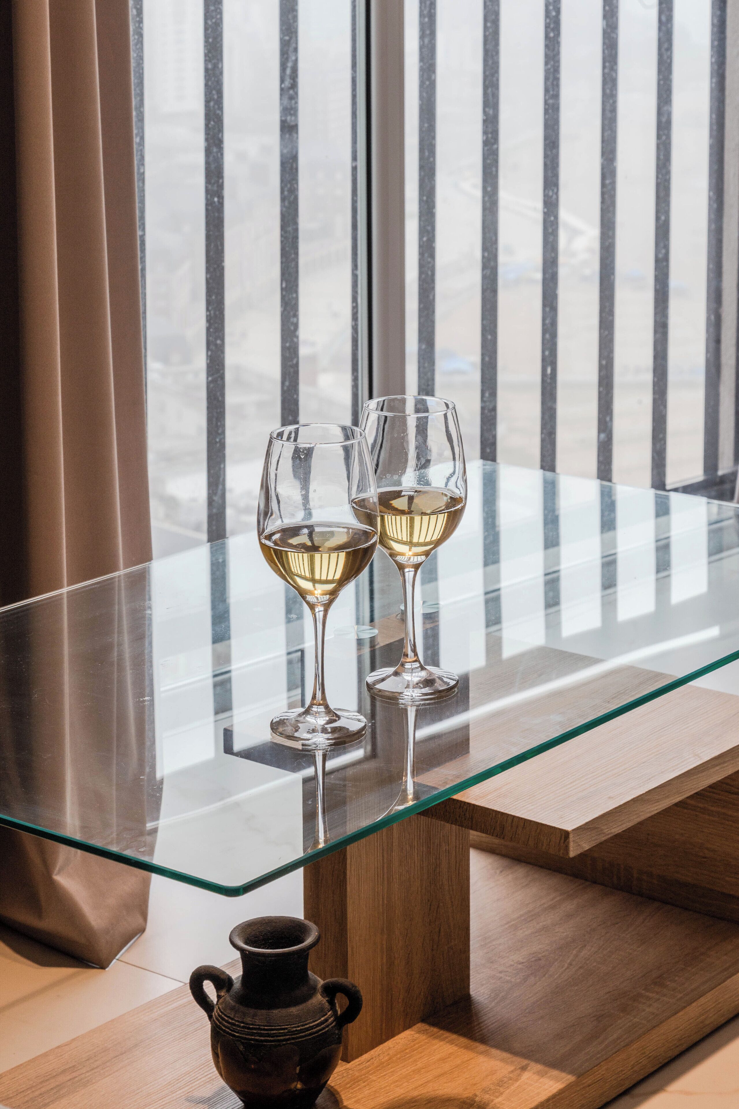 Una mesa de cristal con dos copas de vino, un florero y una vista a través de una ventana con persianas verticales.