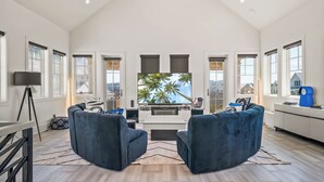 A spacious living room with a large TV, two blue sofas, and a view of palm trees through the windows. - 160 COA Dune Or Do Not (Port Aransas)