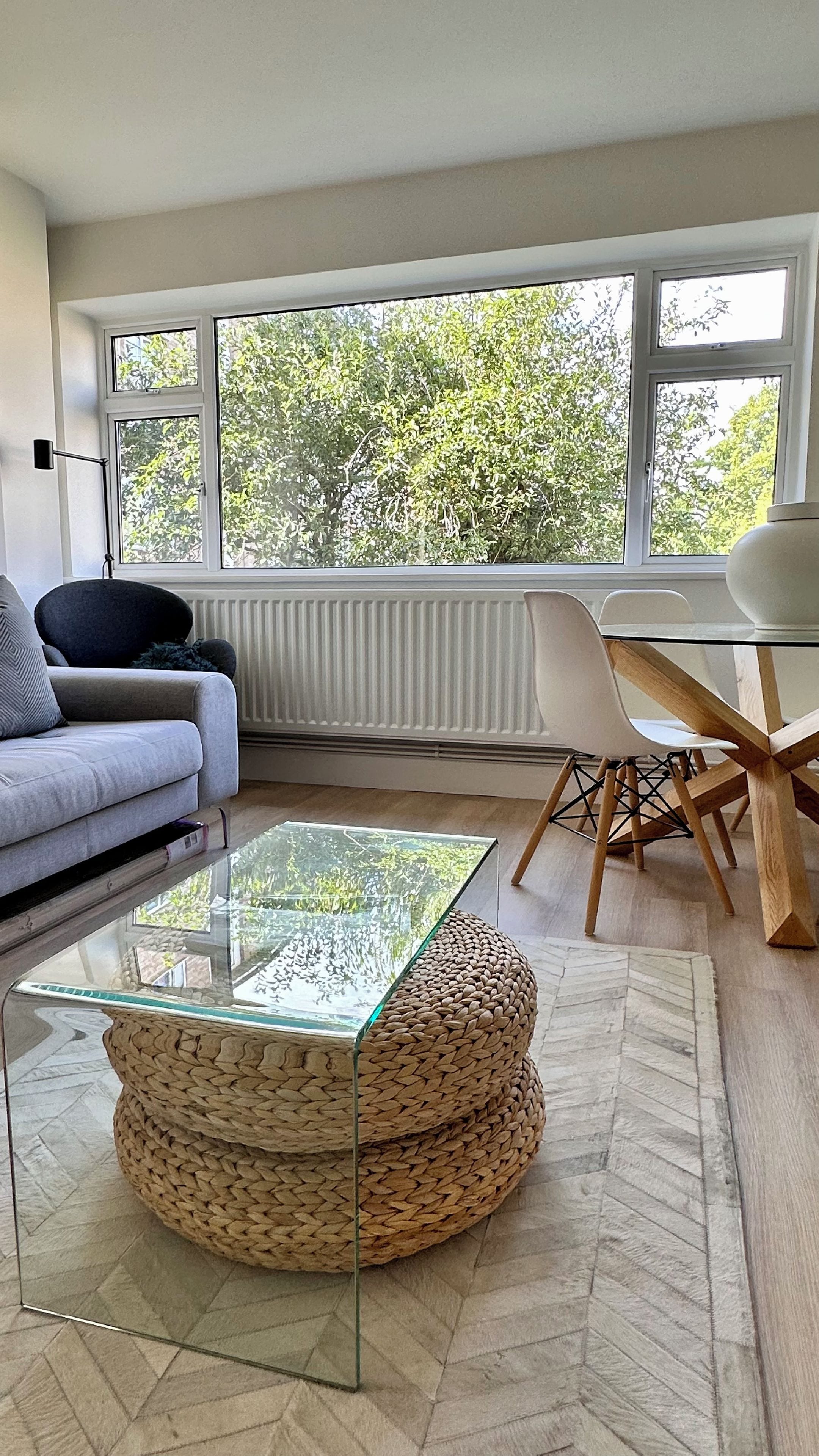 A modern living room with a glass coffee table, a sofa, a dining table with chairs, and a large window with a view of trees.