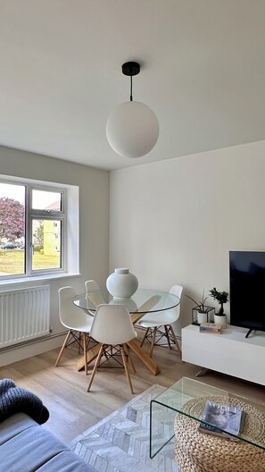 Ein modernes Wohnzimmer mit einem Glastisch, weißen Stühlen, einer Couch, einem Fernseher und einer Pendelleuchte.