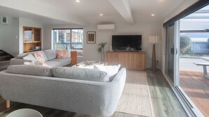 A modern living room with a grey sofa, a flat-screen TV, and a large sliding glass door leading to an outdoor area. - Absolute Lakefront | Panoramic Views & Spacious Stay for 10 (Rotorua)