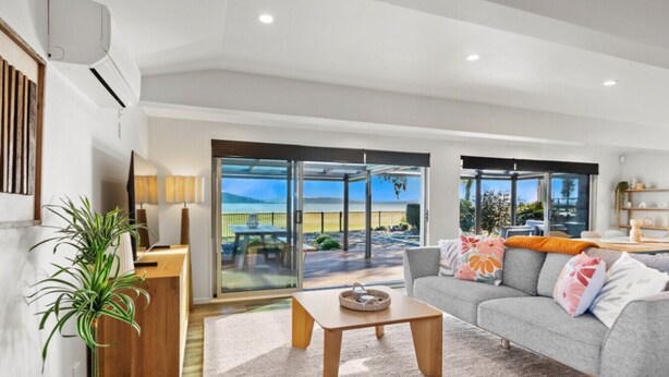 A modern living room with a grey sofa, a wooden coffee table, and a large sliding glass door leading to an outdoor patio. - Absolute Lakefront | Panoramic Views & Spacious Stay for 10 (Rotorua)