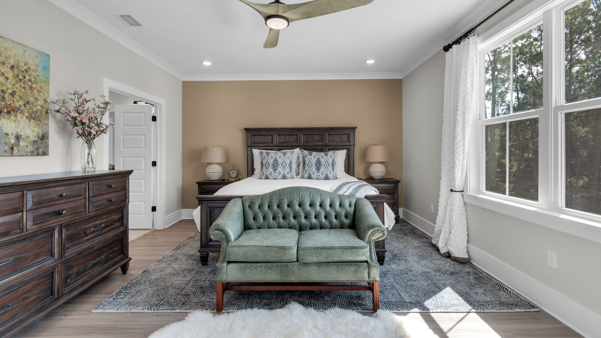 A bedroom with a large bed, a ceiling fan, a dresser, and a window with curtains.