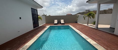 A rectangular swimming pool with a wooden deck surrounding it, two lounge chairs facing the pool, and a clear sky above.