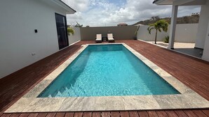 Una piscina rectangular con una cubierta de madera que la rodea, dos sillas de descanso y vistas a un paisaje montañoso bajo un cielo parcialmente nublado.