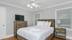 Une chambre à coucher bien rangée, avec un lit, un téléviseur posé sur une commode en bois, une table de chevet avec une lampe et un ventilateur de plafond.