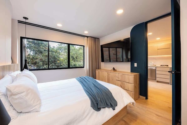 A modern bedroom with a large bed, a wooden dresser, and a view of the outdoors through a large window.