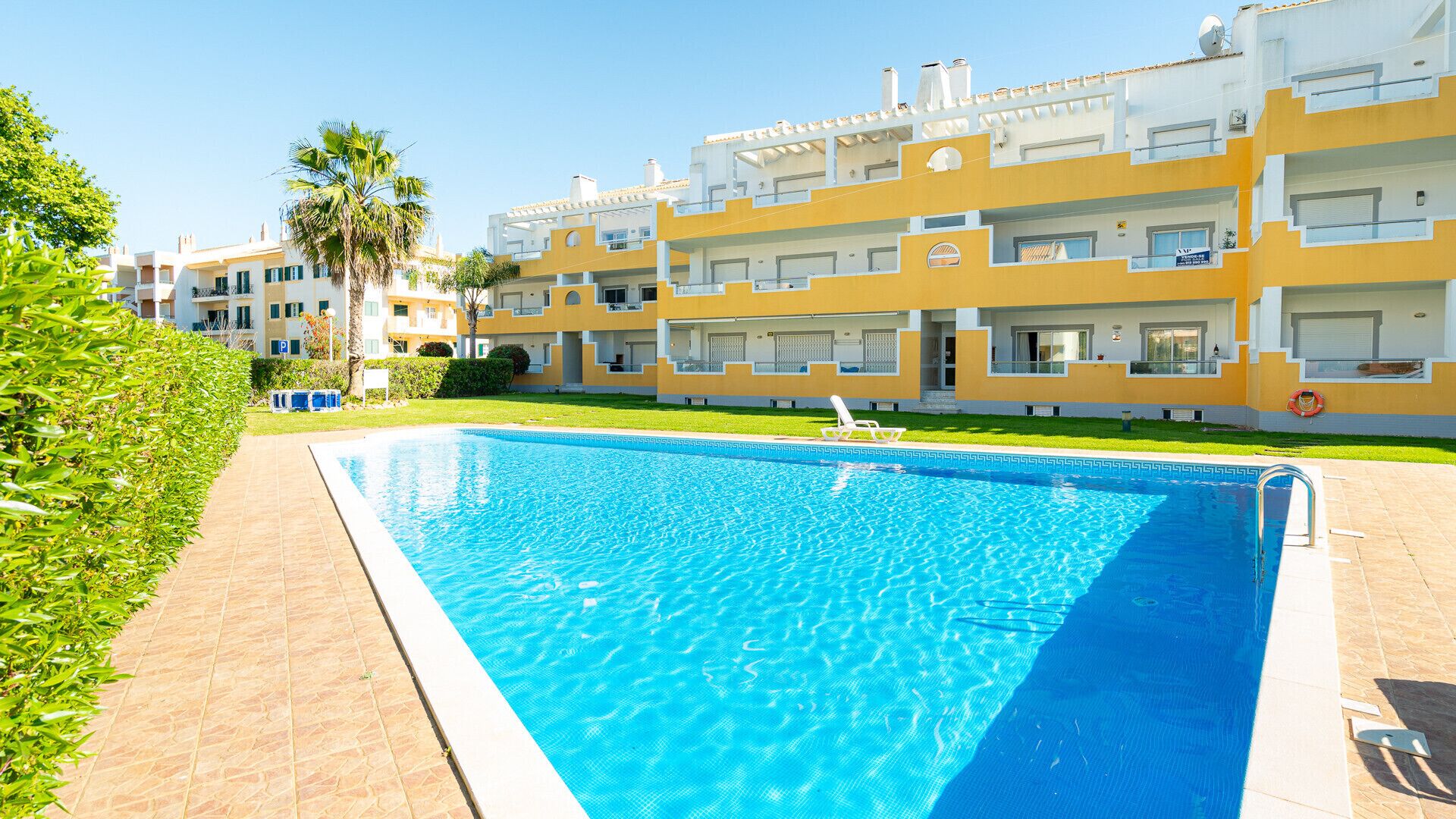 Una piscina con solárium empedrado, rodeada de césped cuidado y una hilera de edificios de varios pisos con balcones.