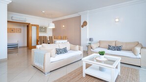A modern living room with two beige sofas, a white coffee table, and a dining area with striped chairs. - FALESIA MARINA APARTMENT (Vilamoura)