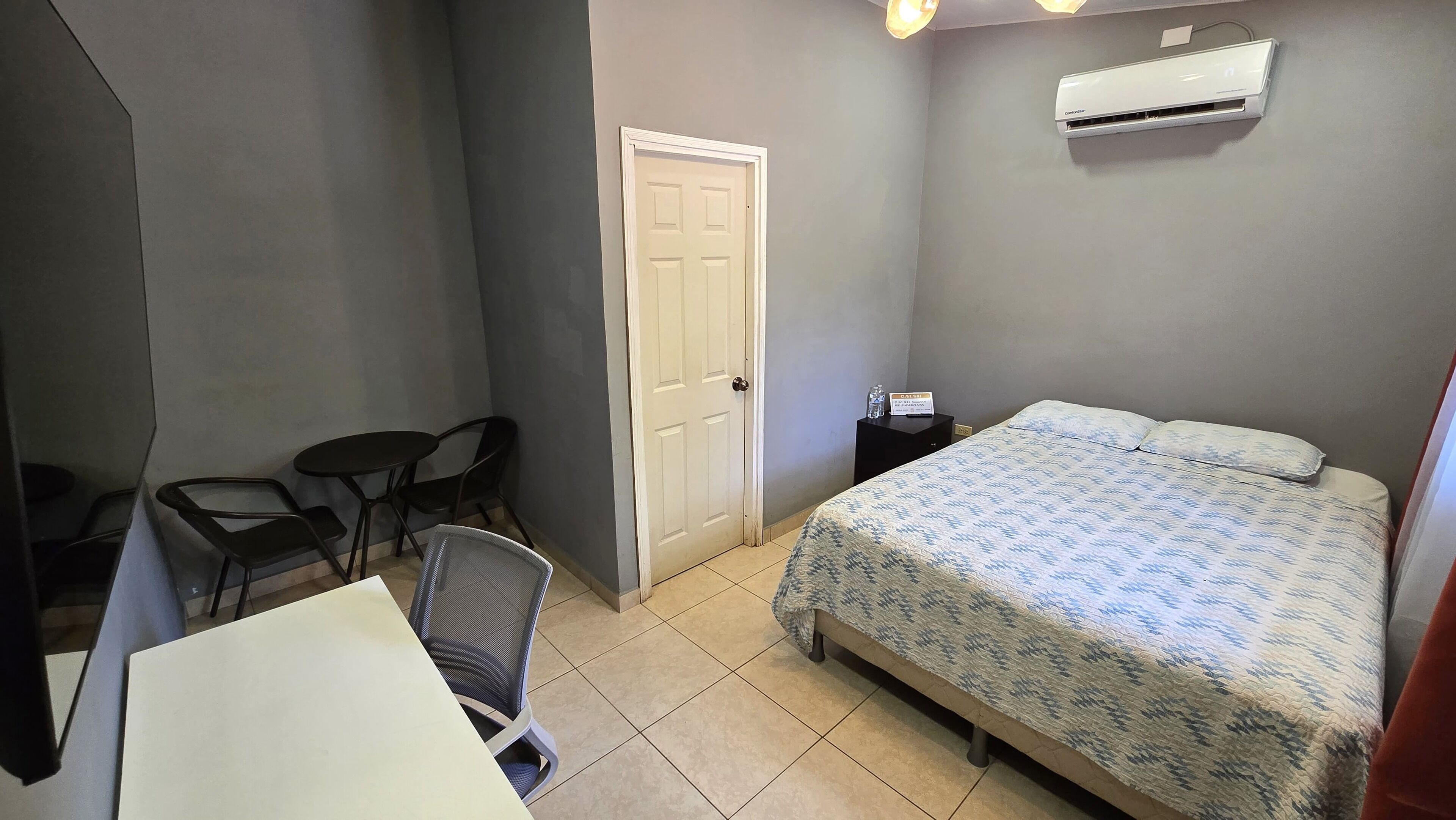 Een kleine, verzorgde slaapkamer met een bed, een bureau, twee stoelen, een deur en een airconditioning.