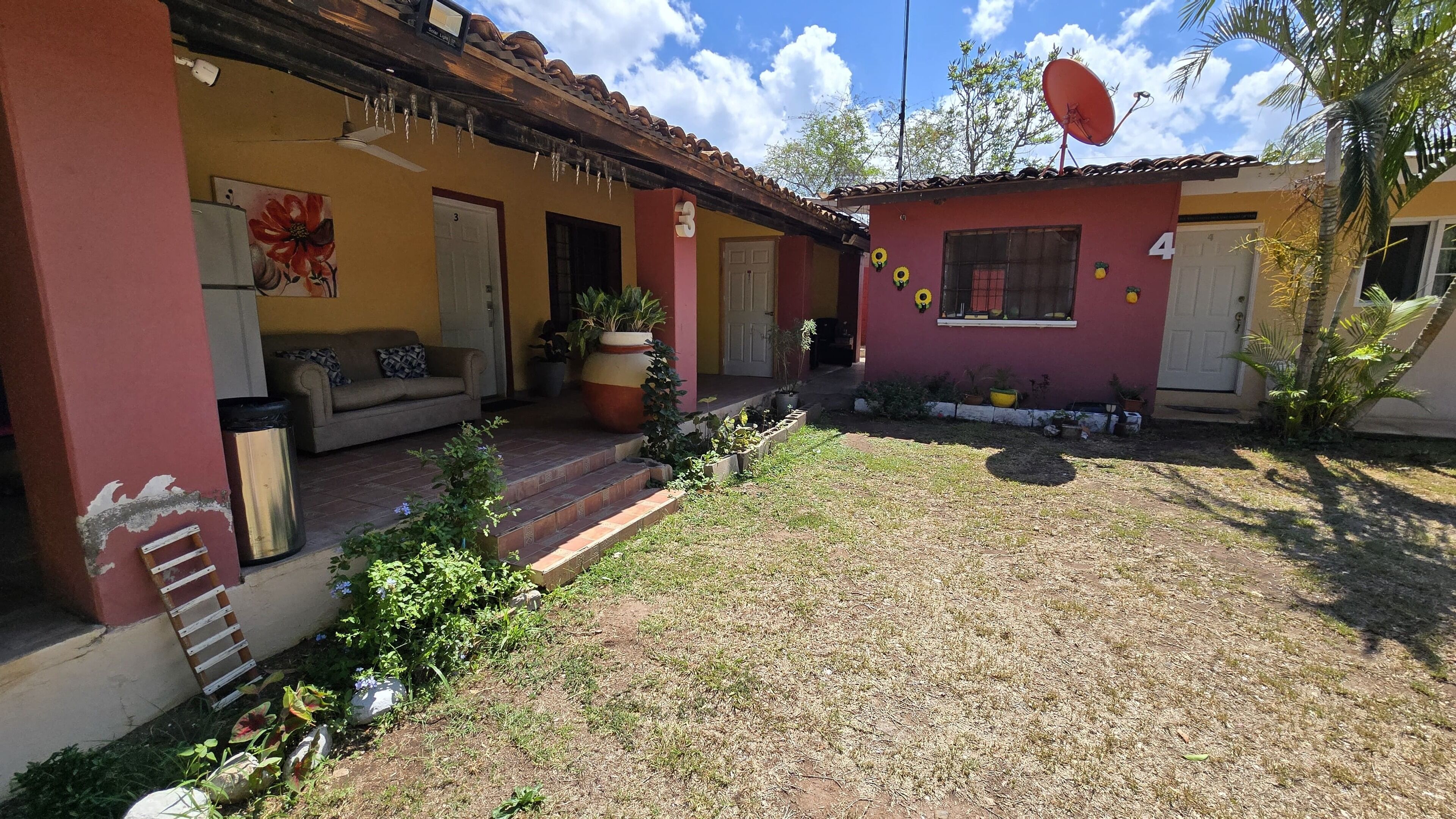 Un edificio de dos pisos con fachada roja y amarilla, puertas numeradas y un pequeño jardín con plantas en macetas y un banco.