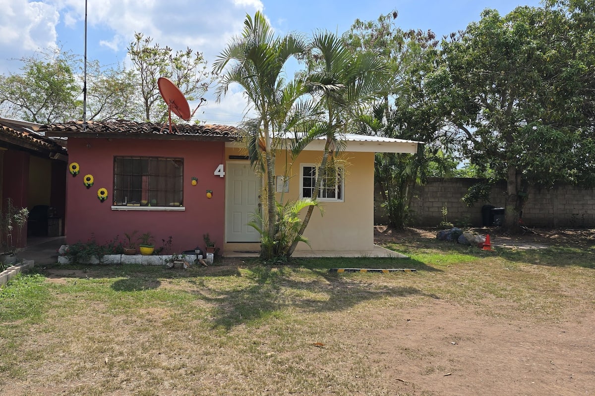 Ein zweigeschossiges Haus mit roter und gelber Fassade, einer Satellitenschüssel und einem kleinen Garten mit Topfpflanzen und einer Palme.