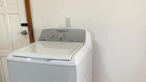 Une machine à laver à chargement frontal blanche, dans une pièce dont le sol est recouvert de parquet et qui possède une porte blanche.