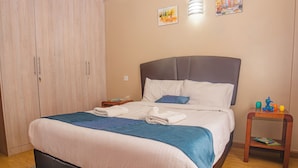 Cama bem arrumada com edredom azul e branco, mesa de cabeceira de madeira com luminária e guarda-roupa de madeira.