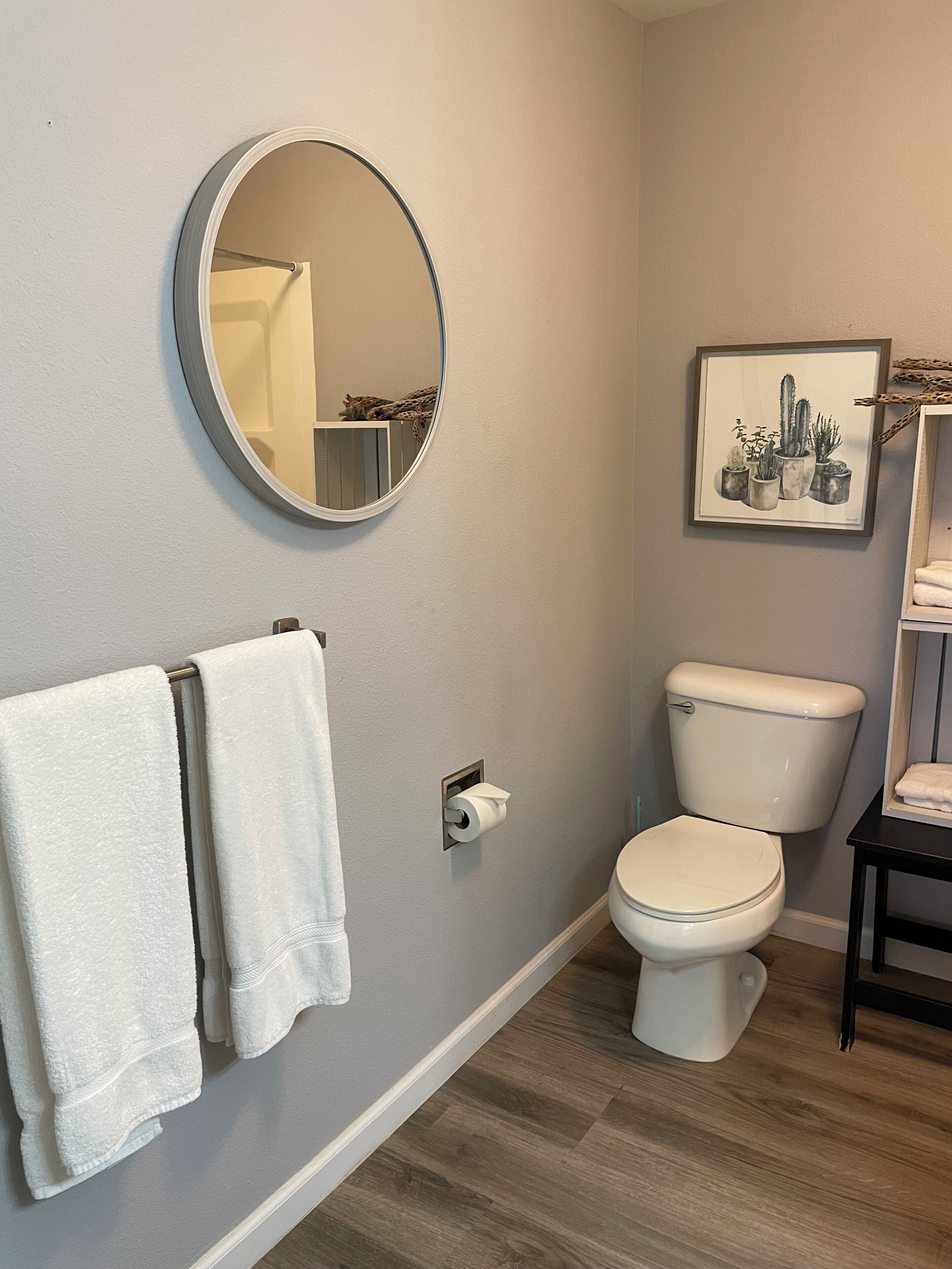 A bathroom with a toilet, a towel rack with two towels, a toilet paper holder, a framed picture of plants, and a mirror.