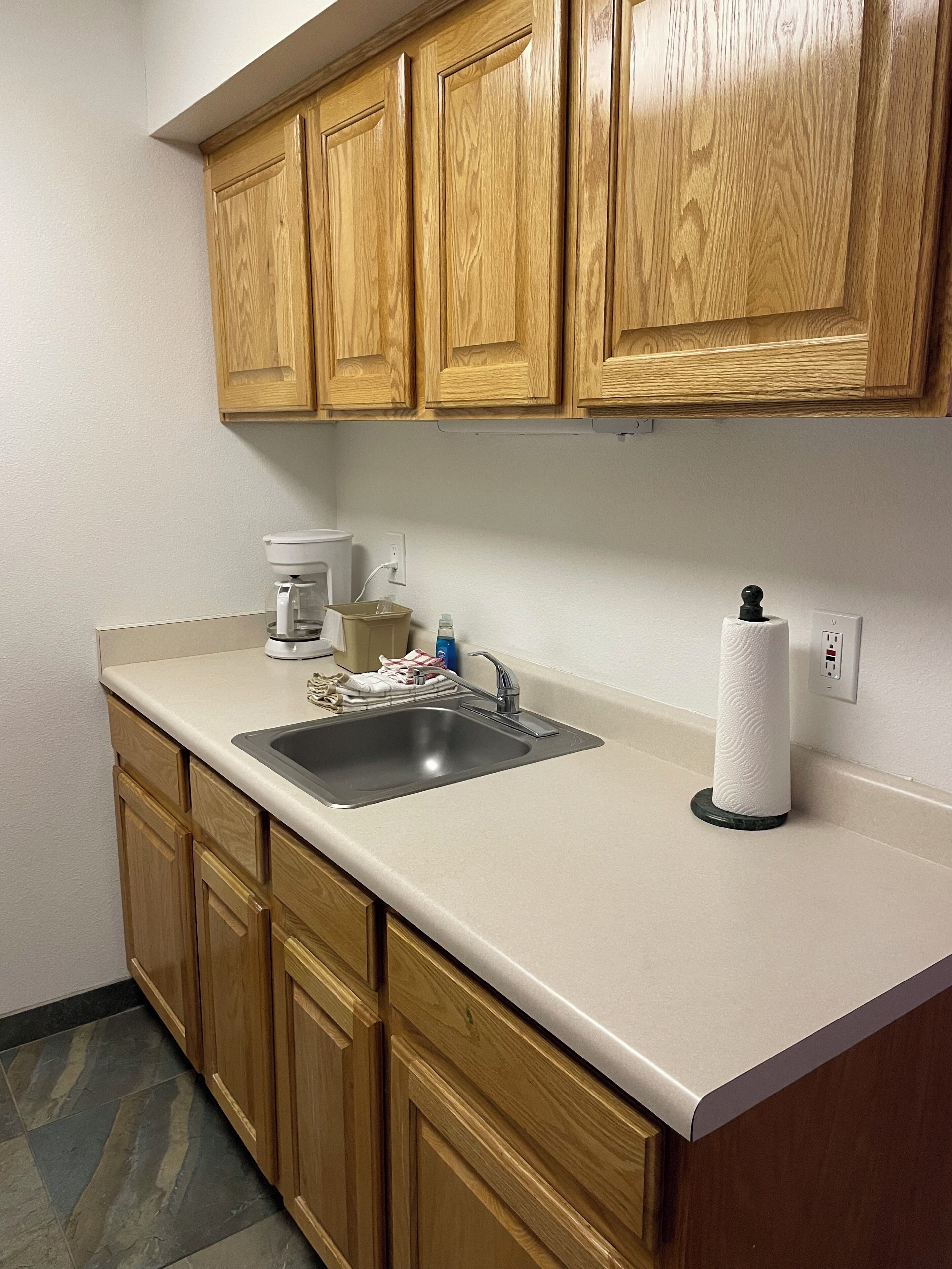 A kitchen with wooden cabinets, a stainless steel sink, a coffee maker, and a paper towel dispenser.