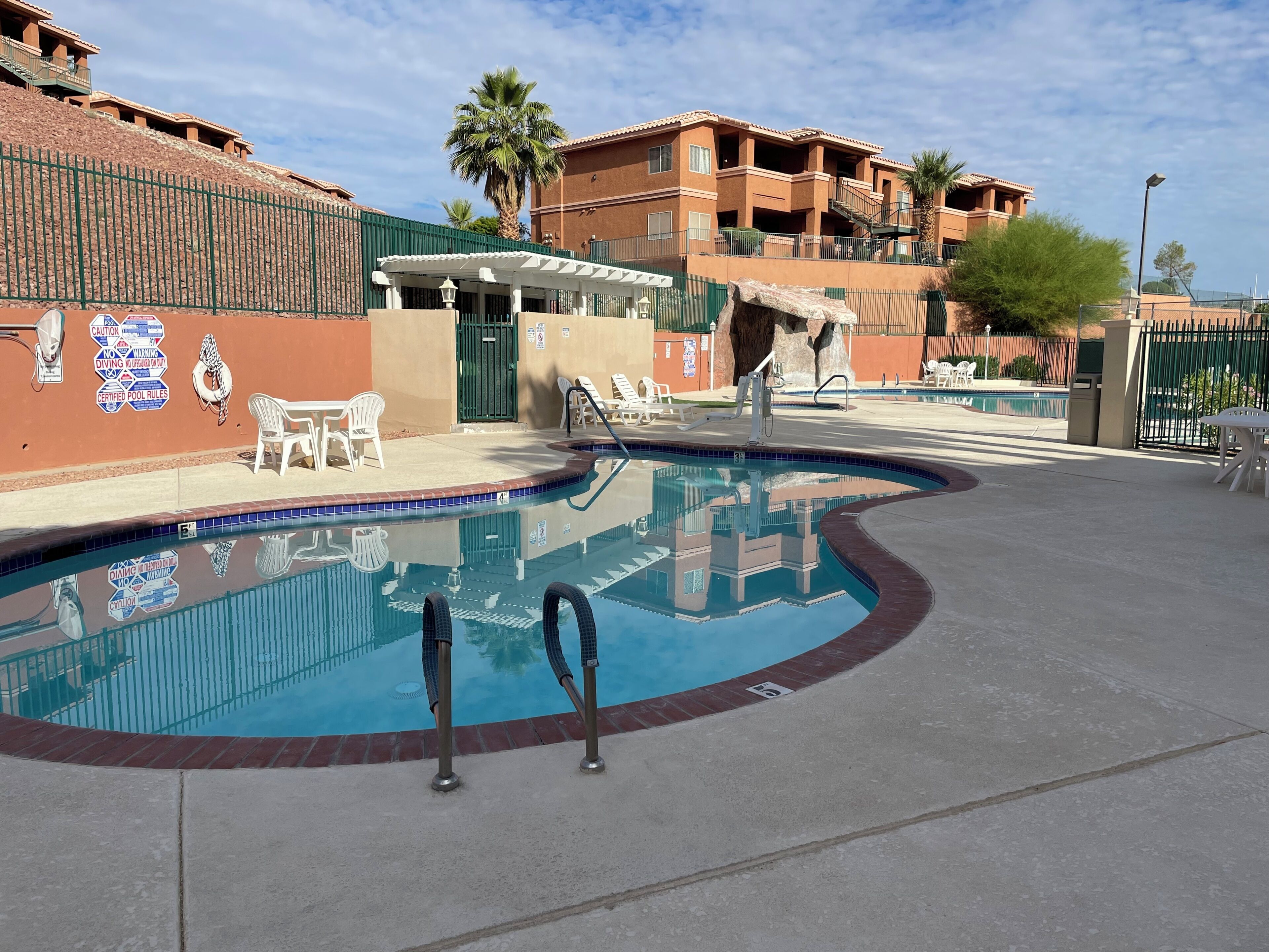 A swimming pool with lounge chairs, a covered seating area, and a building with balconies in the background.