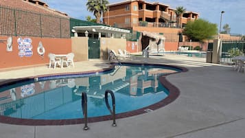 A swimming pool with lounge chairs, a covered seating area, and a building with balconies in the background.