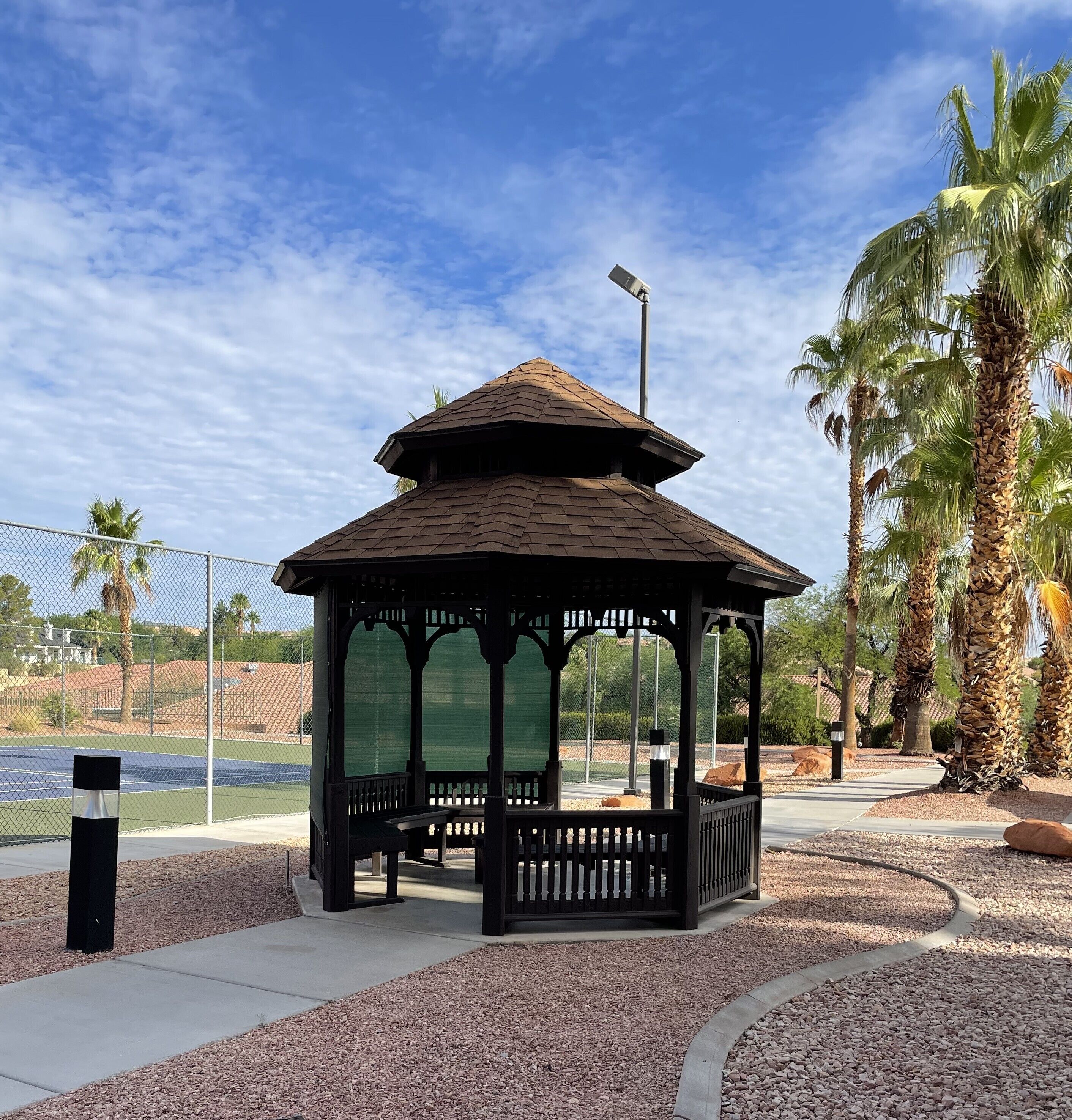 A gazebo with a peaked roof, surrounded by gravel and palm trees, with a tennis court and fence in the background.