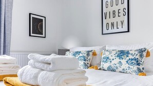 Un lit bien fait, avec des serviettes blanches et jaunes, des coussins à motifs floraux et une affiche sur le mur avec l’inscription « Seules les bonnes ondes sont les bienvenues ».