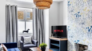 Un salon moderne avec un canapé, un fauteuil et une table basse. Un téléviseur est fixé au mur, et il y a une fenêtre avec des rideaux.