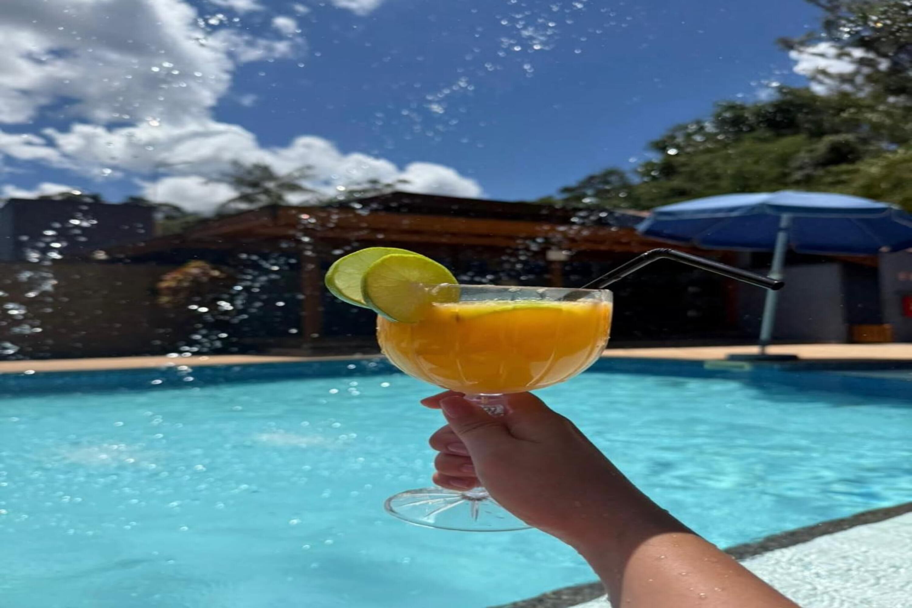 Eine Hand hält einen Cocktail mit einer Limettenscheibe, im Hintergrund ein Pool und ein Gebäude.