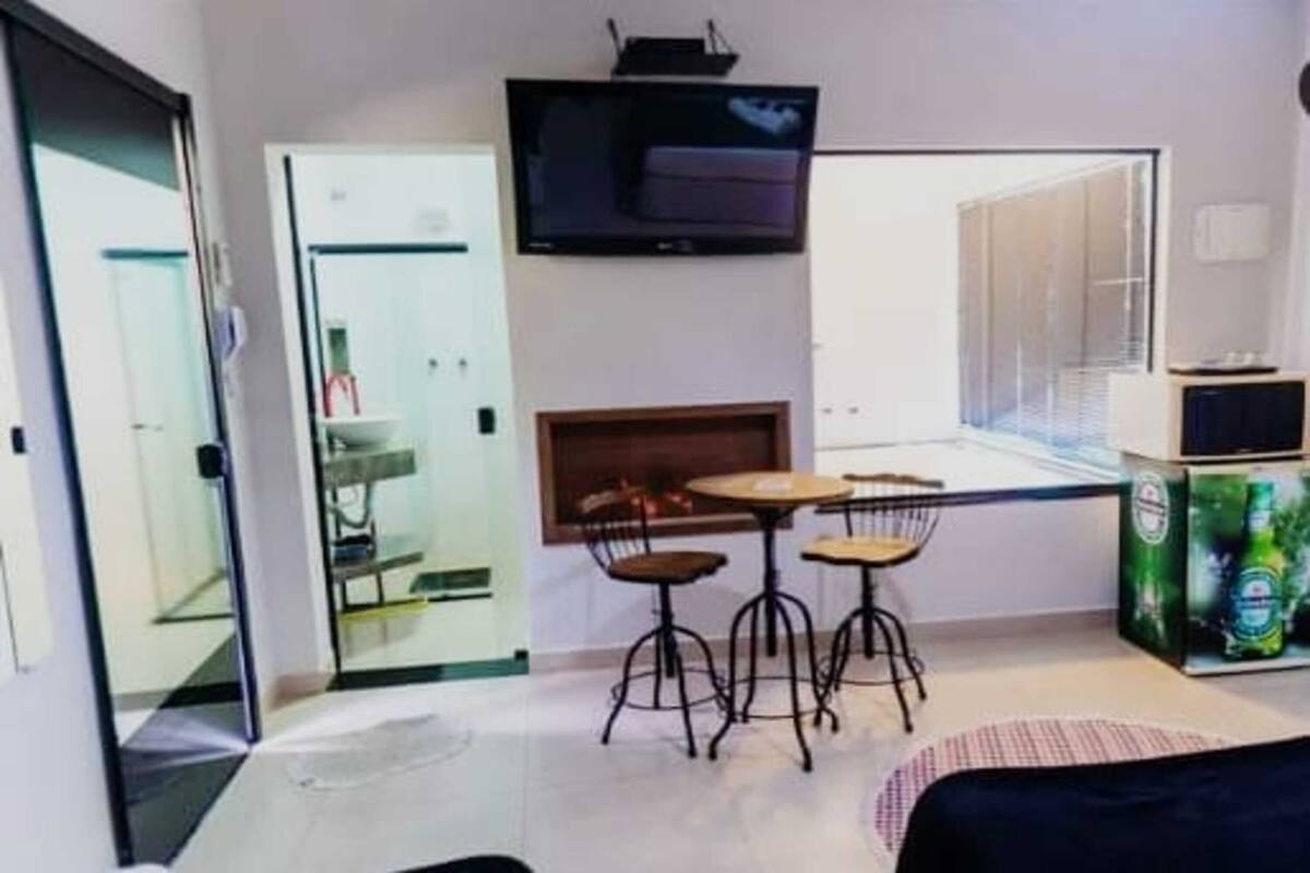 Un espacio habitable compacto con una puerta de vidrio que da a un baño, un pequeño comedor con taburetes, un televisor de pantalla plana montado en la pared y un estante con bebidas embotelladas.