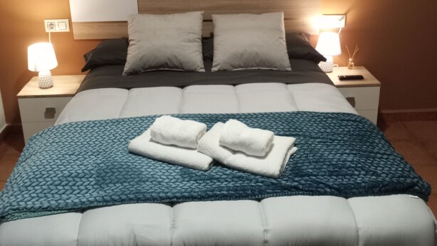 A neatly made bed with a blue quilt, two pillows, and two folded towels on the bed. There are two bedside tables with lamps on either side of the bed.