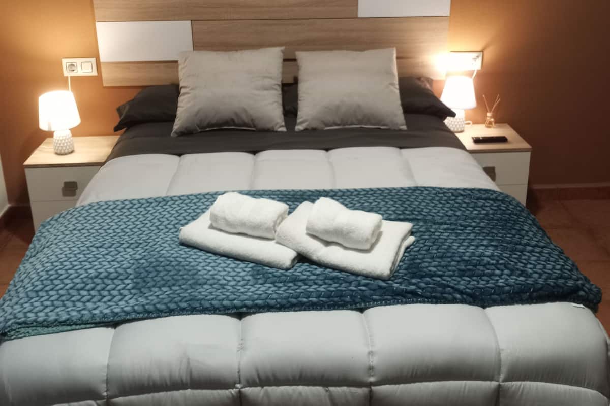 Una cama bien hecha con un edredón azul, dos almohadas y dos toallas dobladas. Hay dos mesitas de noche con lámparas a cada lado de la cama.
