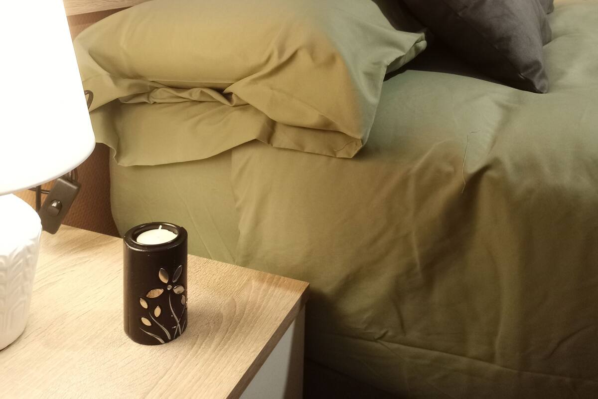 Una cama bien hecha con ropa de cama verde y dos almohadas, una mesita de noche con una lámpara y una vela, y una pared decorada con una amapola roja.