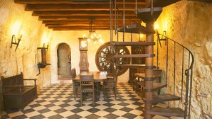 Ein Zimmer mit Schachbrettmuster-Boden, einem Holztisch mit Stühlen, einer großen Holztreppe im Wendelgang und Steinwänden.