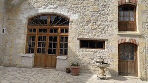 Ein Steinhaus mit einem großen Bogenfenster, einem kleineren Fenster und einer Tür. Vor der Fassade stehen Kübelpflanzen und eine Zierteiche.