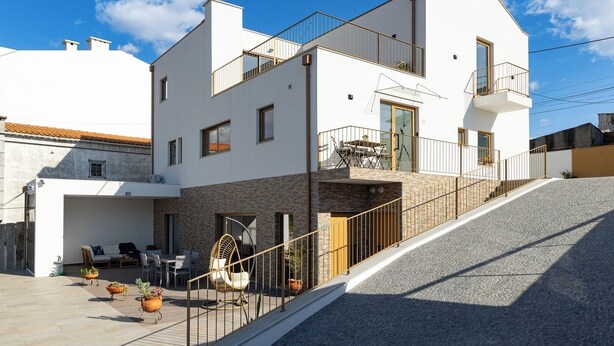 A modern two-story house with a balcony, a stone facade, and a paved patio area with outdoor furniture and potted plants.