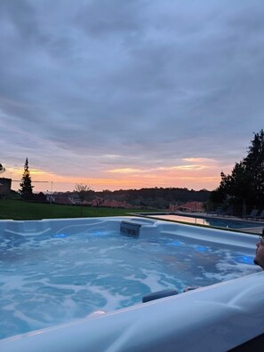 A hot tub with a view of a sunset and a grassy area with trees and buildings in the background.