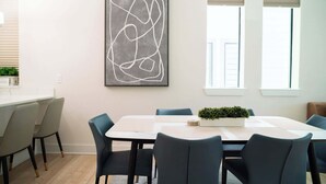 A modern dining area with a white table, dark chairs, a geometric art piece, and a window with blinds.
