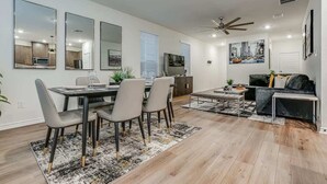 A modern living room with a dining area, a ceiling fan, and a large window with blinds.